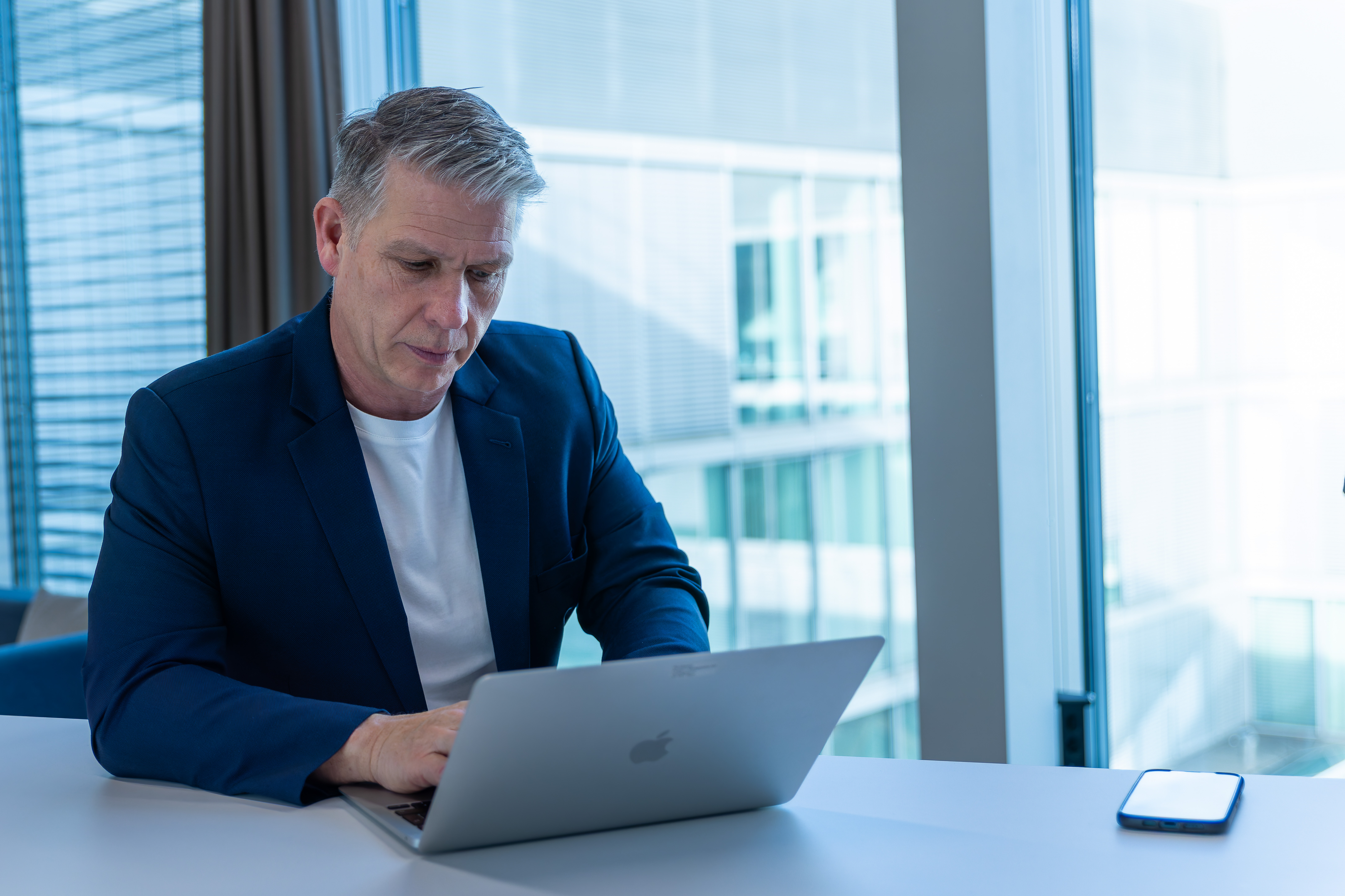 Erfahrener Business Mann in Jacket schaut zufrieden lächelnd in die Kamera, Ihr Team verschwommen hinter Ihr in den Büros von Headhunter Bern und Personalberatung Bern.