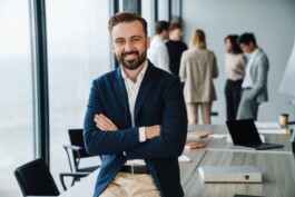 Berater von SAP Bern schaut freundlich, lächelnd direkt in die Kamera, ist angelehnt an Sitzungstisch, seine Kollegen sind im Hintergrund im Sitzungszimmer 